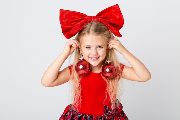 Una linda niña con un vestido rojo y un lazo en la cabeza sosteniendo juguetes para árboles de Navidad. fondo blanco, espacio para texto