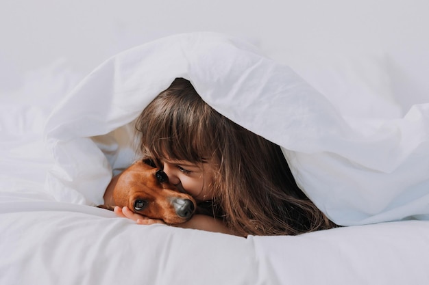 linda niñita morena en casa en la cama con un perro dachshund marrón abrazándose y durmiendo. alta calidad