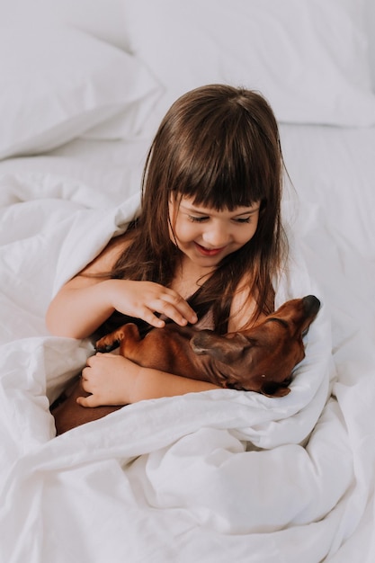 linda niñita morena en casa en la cama con un perro dachshund marrón abrazándose y durmiendo