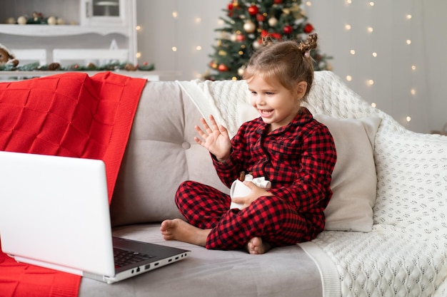 linda niñita en pijama rojo de Navidad usando laptop, teniendo video chat, acostada en el sofá