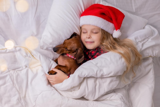 linda niñita rubia con sombrero de Papá Noel y pijamas está durmiendo en la cama con su amado perro mascota