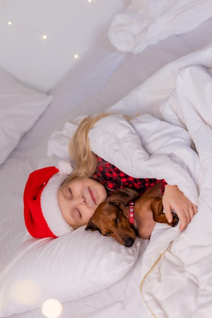 linda niñita rubia con sombrero de Papá Noel y pijamas está durmiendo en la cama con su amado perro mascota