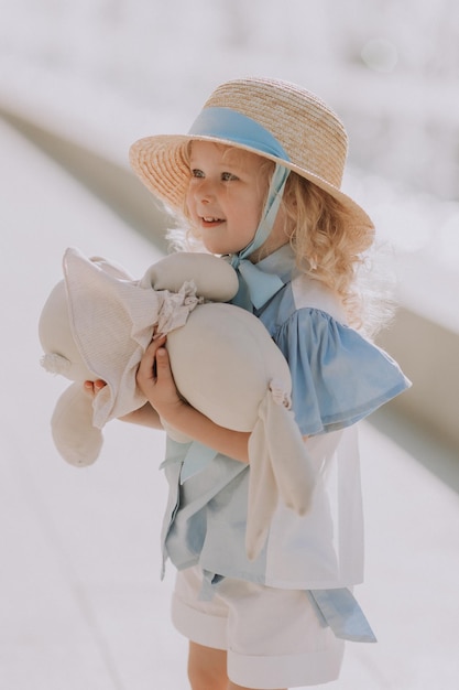 linda niñita rubia con vestido azul y sombrero de paja jugando cerca de la fuente con conejito de peluche, tarjeta
