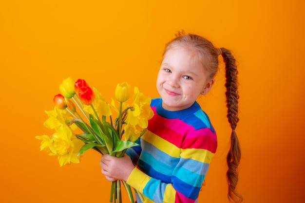 una linda niñita con suéter multicolor sostiene un ramo de flores de primavera con un fondo amarillo escondido