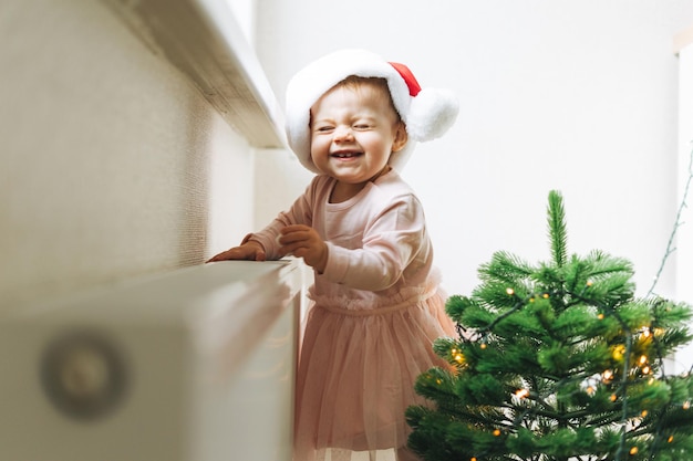 Linda niñita con vestido rosa y gorro de Papá Noel en la habitación con árbol de Navidad en casa feliz Año Nuevo