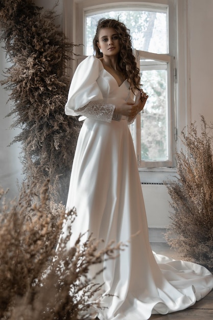 Foto linda noiva caucasiana fica em um vestido de noiva entre flores secas dentro de casa e parece sorrindo para a câmera