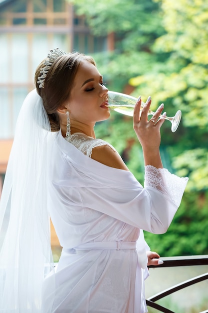Linda noiva com um jaleco branco na varanda bebe champanhe em uma taça