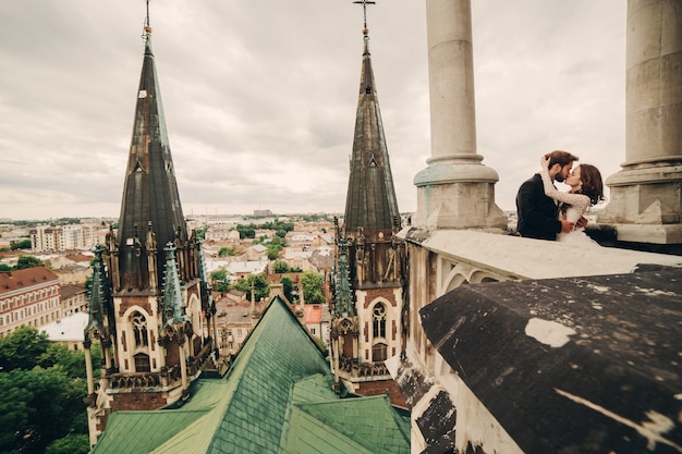 Linda noiva e noivo estiloso se beijando na varanda da antiga catedral gótica com vista panorâmica da cidade