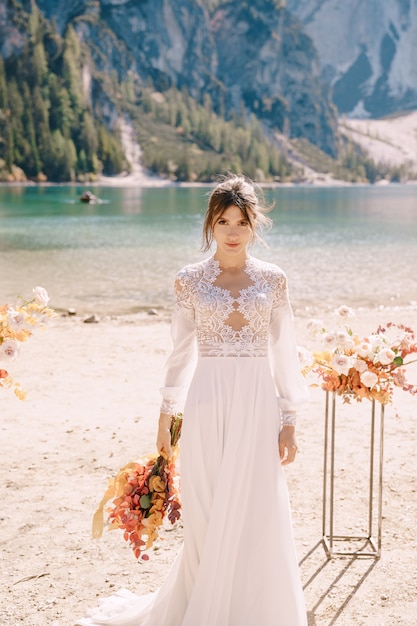 Linda noiva em um vestido branco com mangas e renda, com um buquê amarelo de outono, posando no Lago di Braies, na Itália