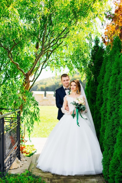 linda noiva em um vestido branco com um longo véu está sorrindo e abraçando com o noivo no parque