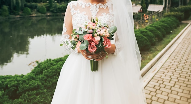 Linda noiva está segurando o buquê nas mãos foto de casamento ao ar livre vestido branco com mangas compridas e renda branca