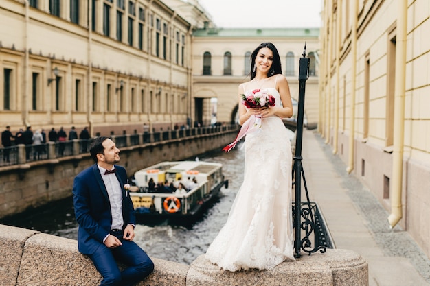 Linda novia adorable con cabello largo y oscuro, usa un vestido de novia, sostiene un hermoso ramo de flores, se para en el puente y su esposo, que se sienta cerca de ella