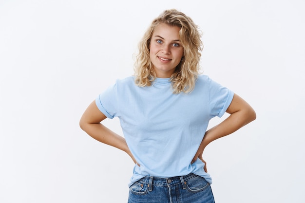 Linda novia esperando a que un amigo se disfrace posando con las manos en la cintura y una sonrisa despreocupada casual, inclinando la cabeza mirando con ojos azules a la cámara sintiéndose relajado y feliz sobre la pared blanca