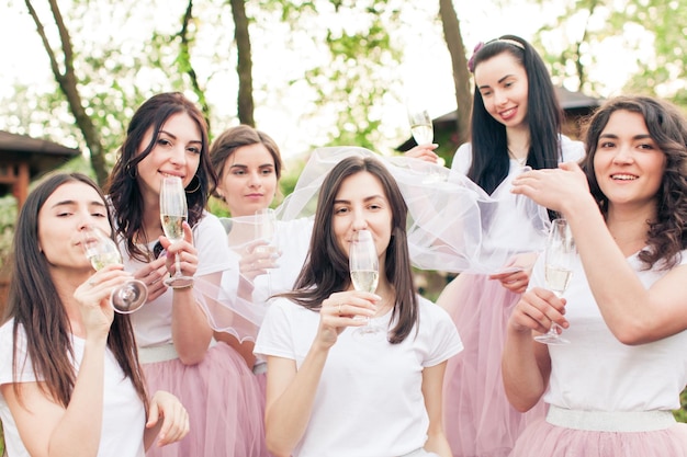 Foto linda novia joven con velo blanco y damas de honor felices a su lado, celebrando la despedida de soltera, bebiendo champán. niñas con velo de novia de la cabeza de su amigo, mirando a la cámara