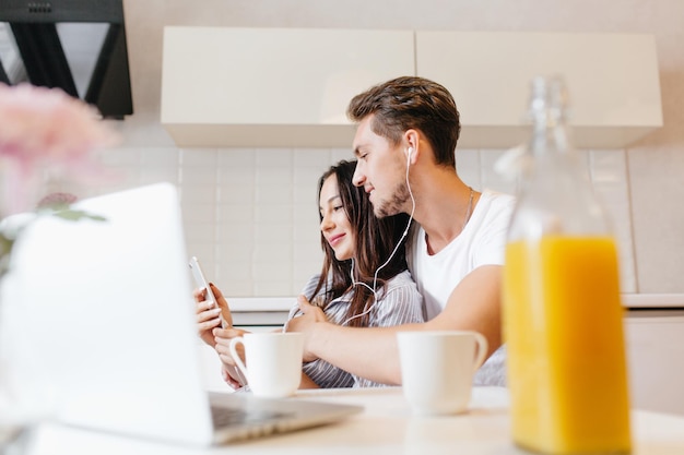 Linda pareja abrazándose mientras usa la computadora portátil en la cocina. Retrato interior de chico escalofriante escuchando música con su novia y disfrutando de un café en una habitación luminosa y acogedora.