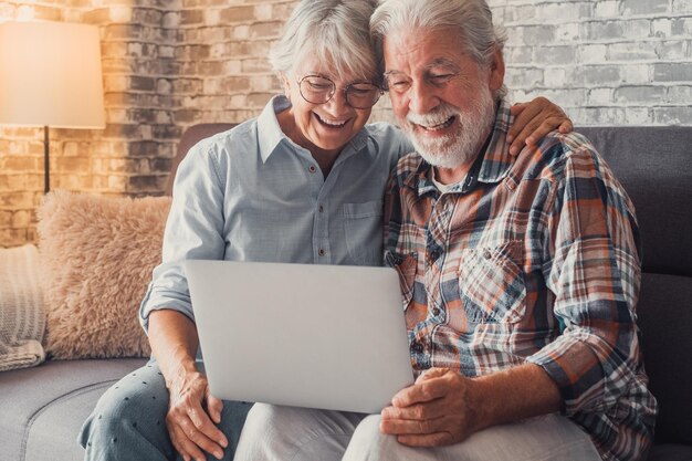 Linda pareja de ancianos sentados en el sofá usando una computadora portátil juntos comprando y navegando por la red Dos personas maduras en la sala de estar disfrutando de la tecnología Retrato de personas mayores riendo enamoradasxA
