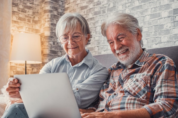 Linda pareja de ancianos sentados en el sofá usando una computadora portátil juntos comprando y navegando por la red Dos personas maduras en la sala de estar disfrutando de la tecnología