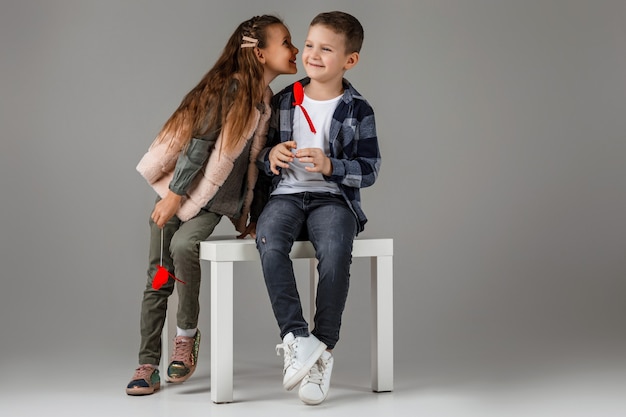 Linda pareja con estilo niña y niño con corazones rojos en palo en ropa de moda sentados juntos