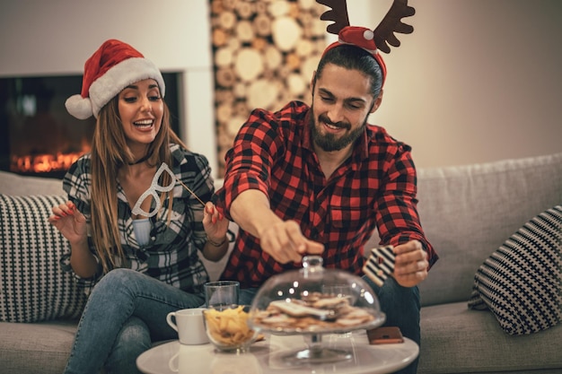 Linda pareja hermosa está pasando la víspera de Año Nuevo en casa sentada en el sofá y comiendo galletas. Pasan unas vacaciones estupendas divirtiéndose en buena compañía en el interior.