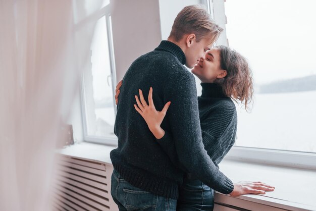 Linda pareja joven abrazándose en el interior cerca de la ventana.