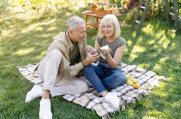 Linda pareja de jubilados haciendo un picnic en el jardín sentados en una manta y comiendo tostadas con mermelada disfrutando