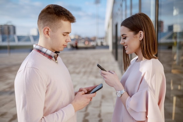 Linda pareja sonriente vestida elegante de pie al aire libre y usando teléfonos inteligentes