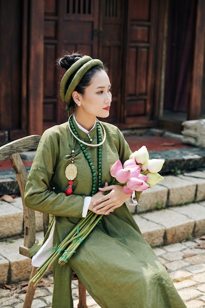 Foto linda pensativa jovem vietnamita com vestido ao dai sentada em um banquinho de madeira com flores de lótus