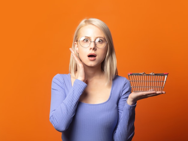 Linda rubia en blusa azul con cesta de la compra sobre un fondo de color naranja exuberante