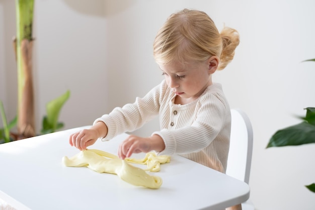 Linda rubia seria de dos años de edad, niña pequeña jugando con limo pegajoso amarillo en el interior de su casa sobre fondo blanco. Concepto de experimento divertido juego desordenado de desarrollo de niños tempranos Espacio de copia