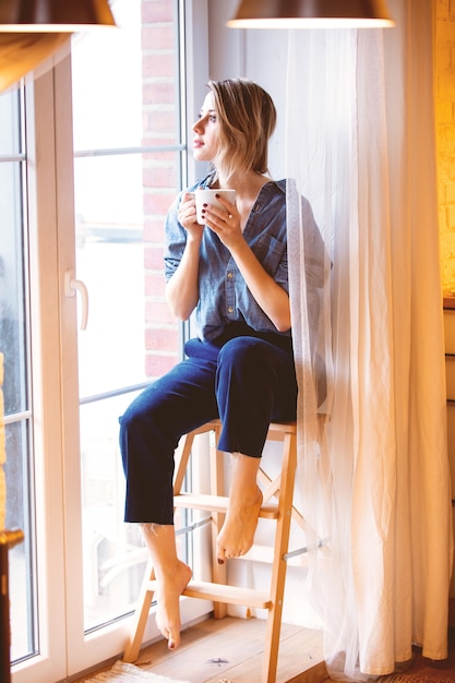 Linda ruiva com uma xícara de café quente sentada na escada de madeira em casa e olhando pela janela