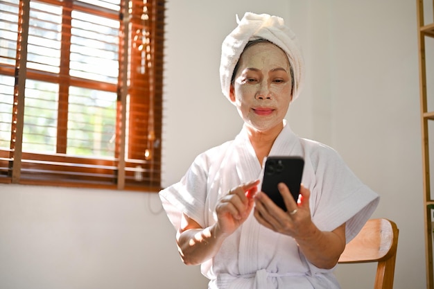 Linda senhora asiática madura em roupão com máscara hidratante facial está usando seu celular