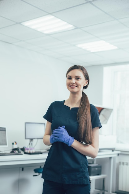 Foto linda senhora de uniforme escuro e luvas de borracha sorrindo e parecendo feliz em pé no laboratório sozinha