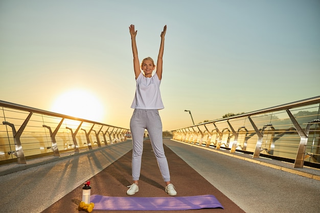 Foto linda senhora desportiva fazendo exercícios e sorrindo em pé na ponte durante o nascer do sol