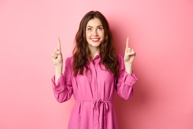 linda senhora feliz no vestido, apontando e olhando para cima e sorrindo alegre, olhando para o texto promocional, lendo seu logotipo, em pé contra a parede rosa.