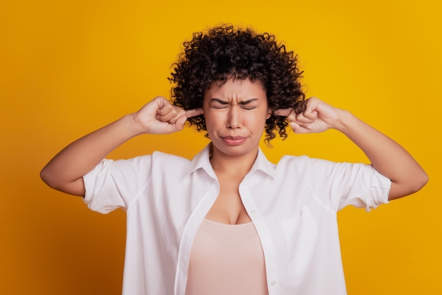 Linda senhora irritada com os dedos tapando as orelhas evitando olhos fechados posando em fundo amarelo