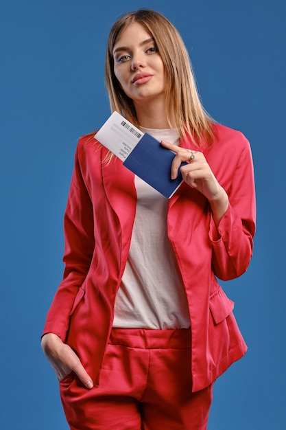 Foto linda senhora loira de blusa branca e terninho vermelho. ela está sorrindo, colocou a mão no bolso, segurando o passaporte e a passagem