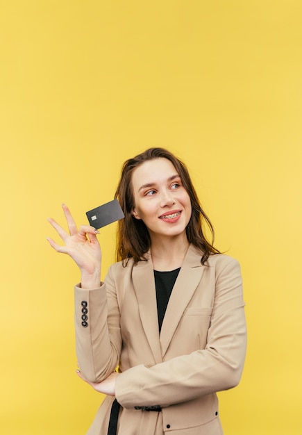 Linda senhora positiva em uma jaqueta bege posa com um cartão bancário em um fundo amarelo e desvia o olhar