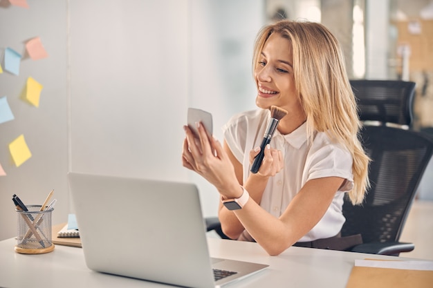 Linda senhora sorridente, aplicando pó com pincel de maquiagem enquanto está sentada à mesa com o laptop no escritório