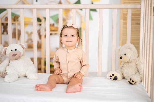 Una linda y sonriente niña sana con traje beige sentada en una cuna en casa en la guardería con osos de peluche en ropa de cama blanca