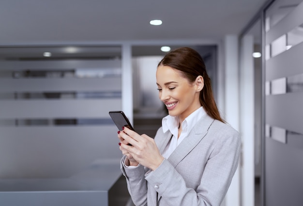 Linda sorridente empresária caucasiana com roupa formal em pé no saguão e usando telefone inteligente.