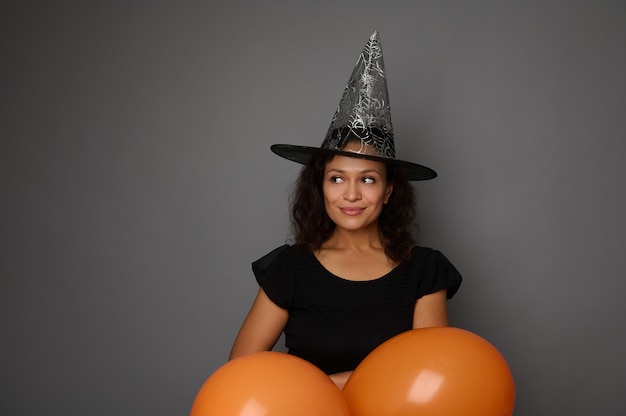 Linda sorridente encantadora mulher latino-americana vestida de preto e chapéu de bruxa espia através dos balões de ar laranja, olha para o lado para um espaço de cópia para anúncio em fundo cinza. Conceito de feliz dia das bruxas