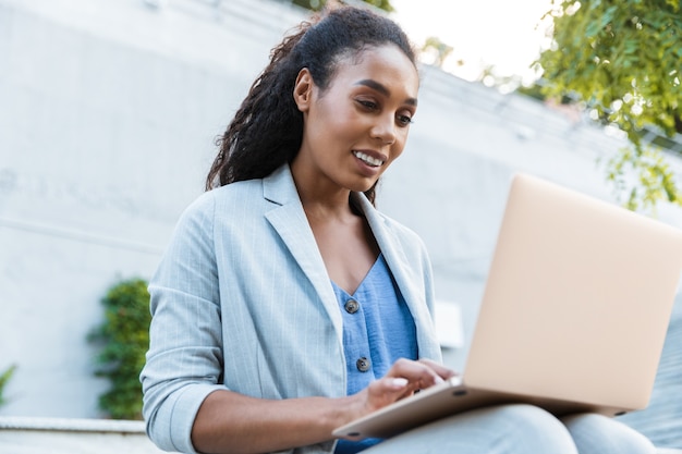 Linda sorridente jovem mulher de negócios africana sentada em um banco ao ar livre, trabalhando em um laptop