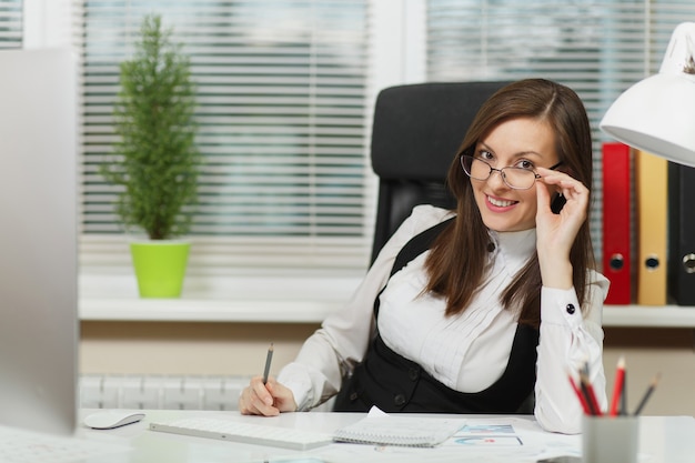 Linda sorridente mulher de negócios de cabelo castanho sexy de terno e óculos, sentada à mesa com uma lâmpada branca em seu local de trabalho, trabalhando no computador com um monitor moderno com documento em um escritório leve