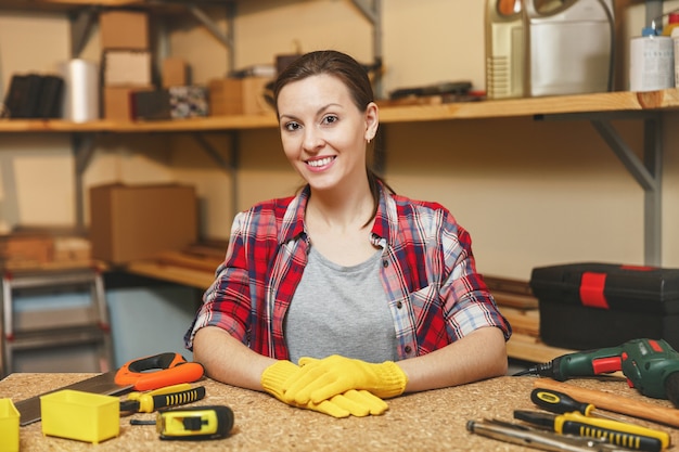 Linda sorridente mulher jovem caucasiana de cabelos castanhos em camisa xadrez, camiseta cinza, luvas amarelas, trabalhando na oficina de carpintaria em uma mesa de madeira local com diferentes ferramentas de trabalho masculinas. Igualdade de gênero.