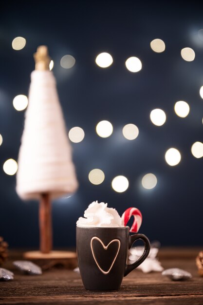 Linda taza pequeña de chocolate caliente con crema batida colmada decorada con caña de azúcar con decoración navideña en el fondo.