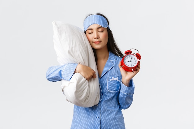 Linda y tonta chica asiática en pijama azul y máscara para dormir, durmiendo en la almohada con los ojos cerrados y mostrando el despertador, teniendo dulces sueños mientras olvidé configurar la alarma, pared blanca