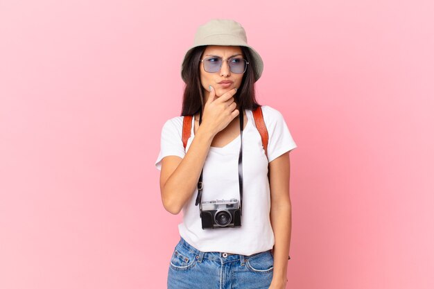 Linda turista hispânica pensando, se sentindo em dúvida e confusa com uma câmera fotográfica e um chapéu