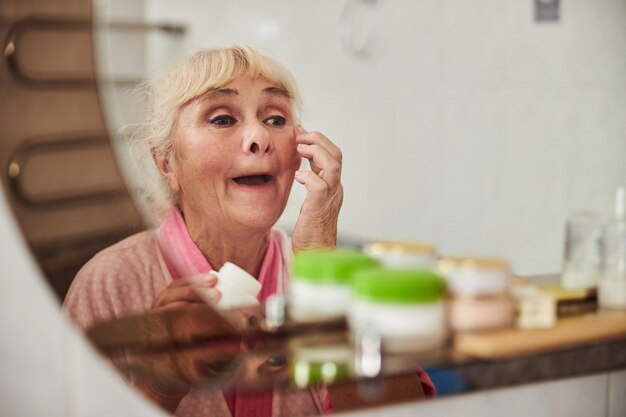 Linda velha aplicando creme no rosto no banheiro