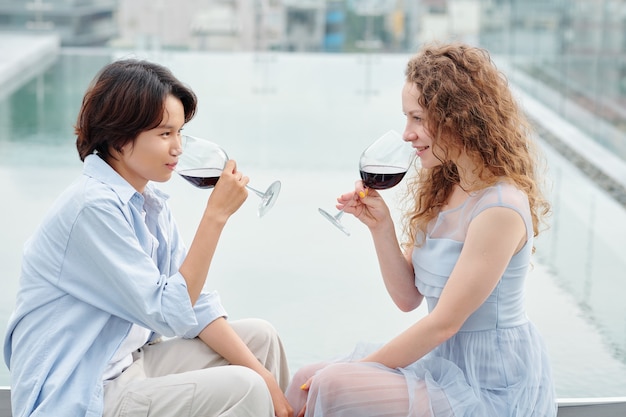 Lindas amigas jovens bebendo vinho tinto e se olhando sentadas no telhado