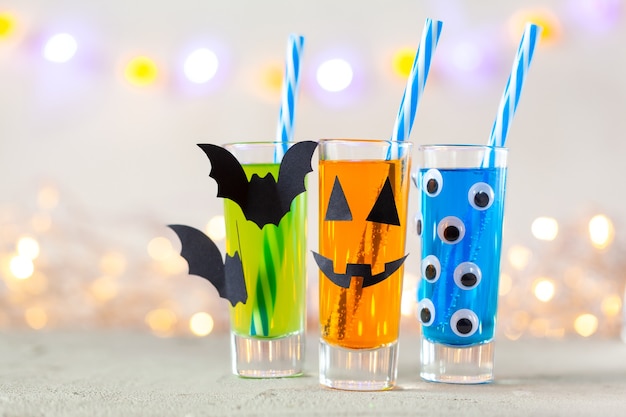 Lindas bebidas de Halloween para una fiesta infantil. Tres coloridos jugo en vidrio, papel de decoración, murciélago y ojos sobre fondo claro con espacio de copia